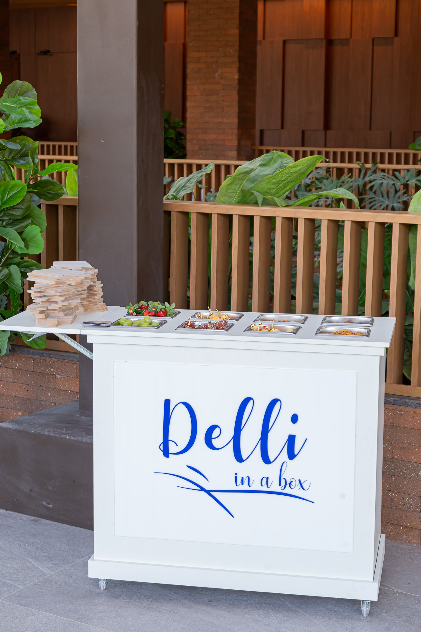 Carrinho Delli in a box branco e elegante, com a logo azul, bandejas de frios e queijos e tábuas de madeira, montado em um evento