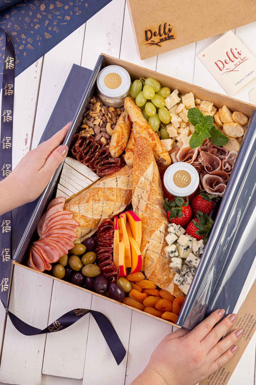 Caixa de frios Delli in a box segurada nas mãos, com fita azul-marinho da marca, kraft com logo, prosciutto, queijos, salame, pães, uvas, morangos, azeitonas e castanhas